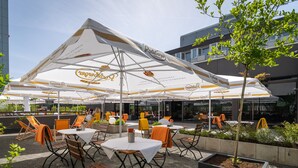 An outdoor patio area with tables and chairs under a large umbrella, surrounded by greenery and buildings. - Best Western Hotel Kaiserslautern (Kaiserslautern)
