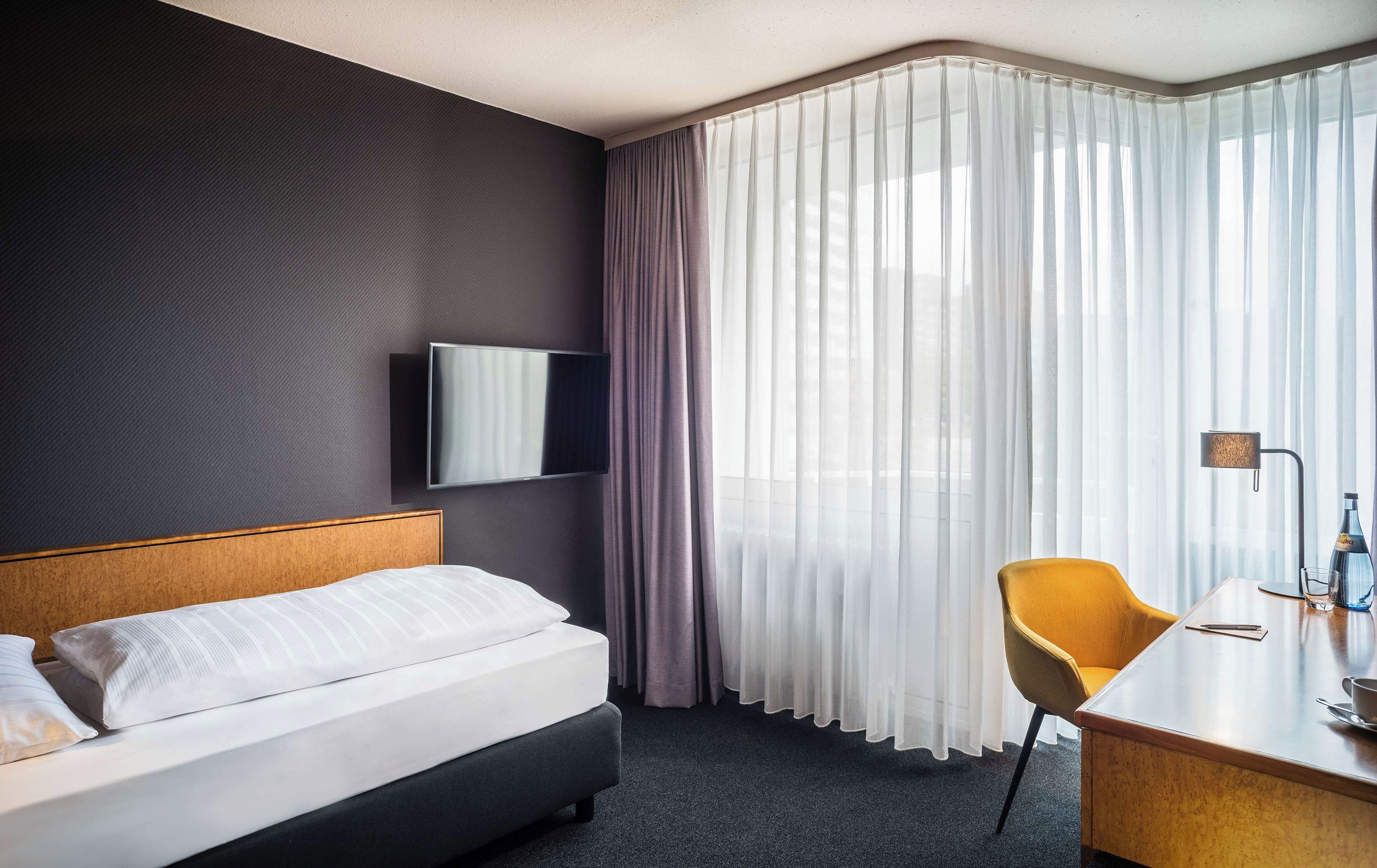 Camera d'albergo con un letto, una scrivania, una sedia, una TV a muro e tende leggere.