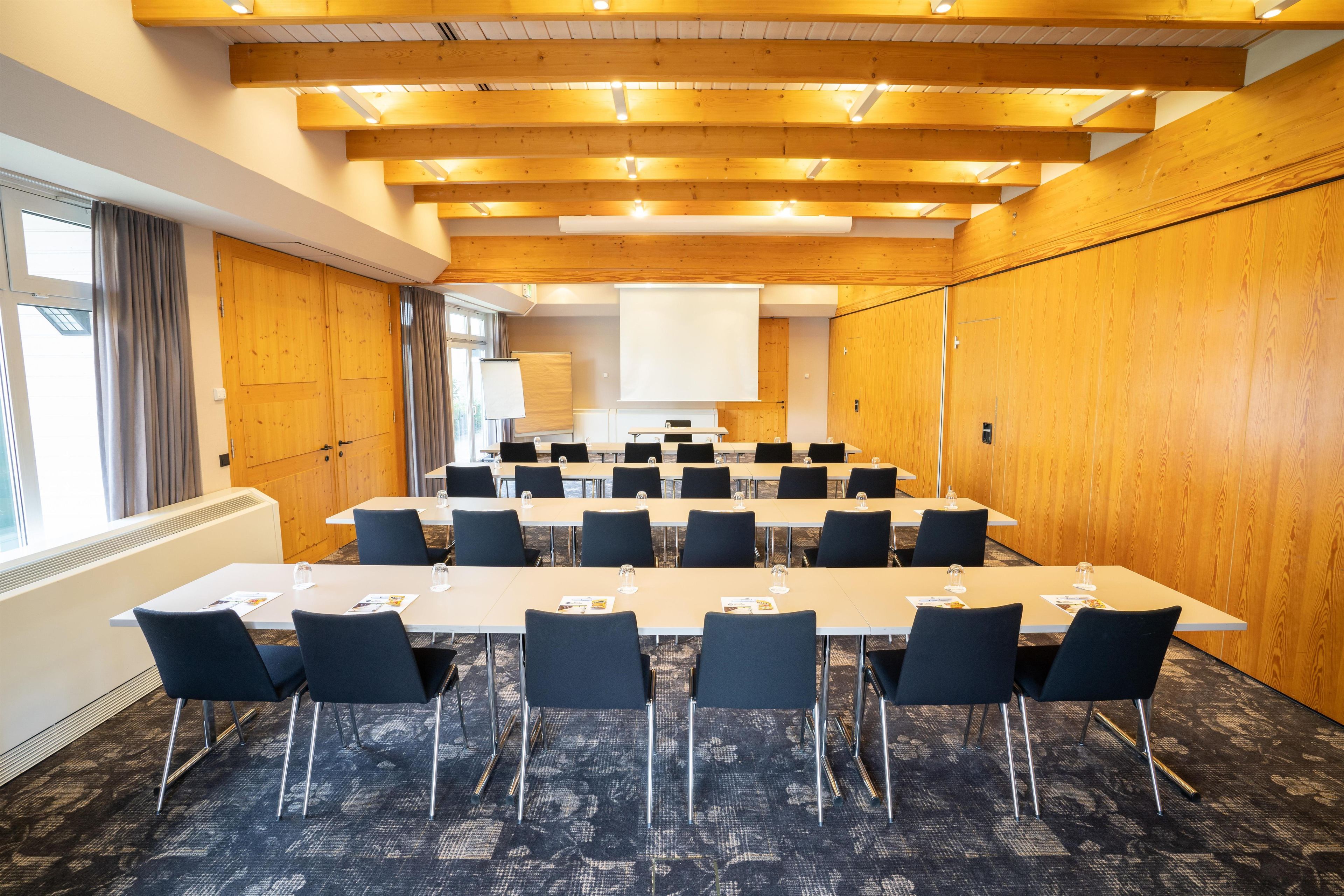 Une salle de conférence avec de longues tables, des chaises, un écran de projection et des panneaux muraux en bois.