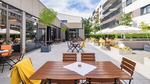 An outdoor dining area with wooden tables and chairs, surrounded by buildings with balconies and outdoor seating. - Best Western Hotel Kaiserslautern (Kaiserslautern)