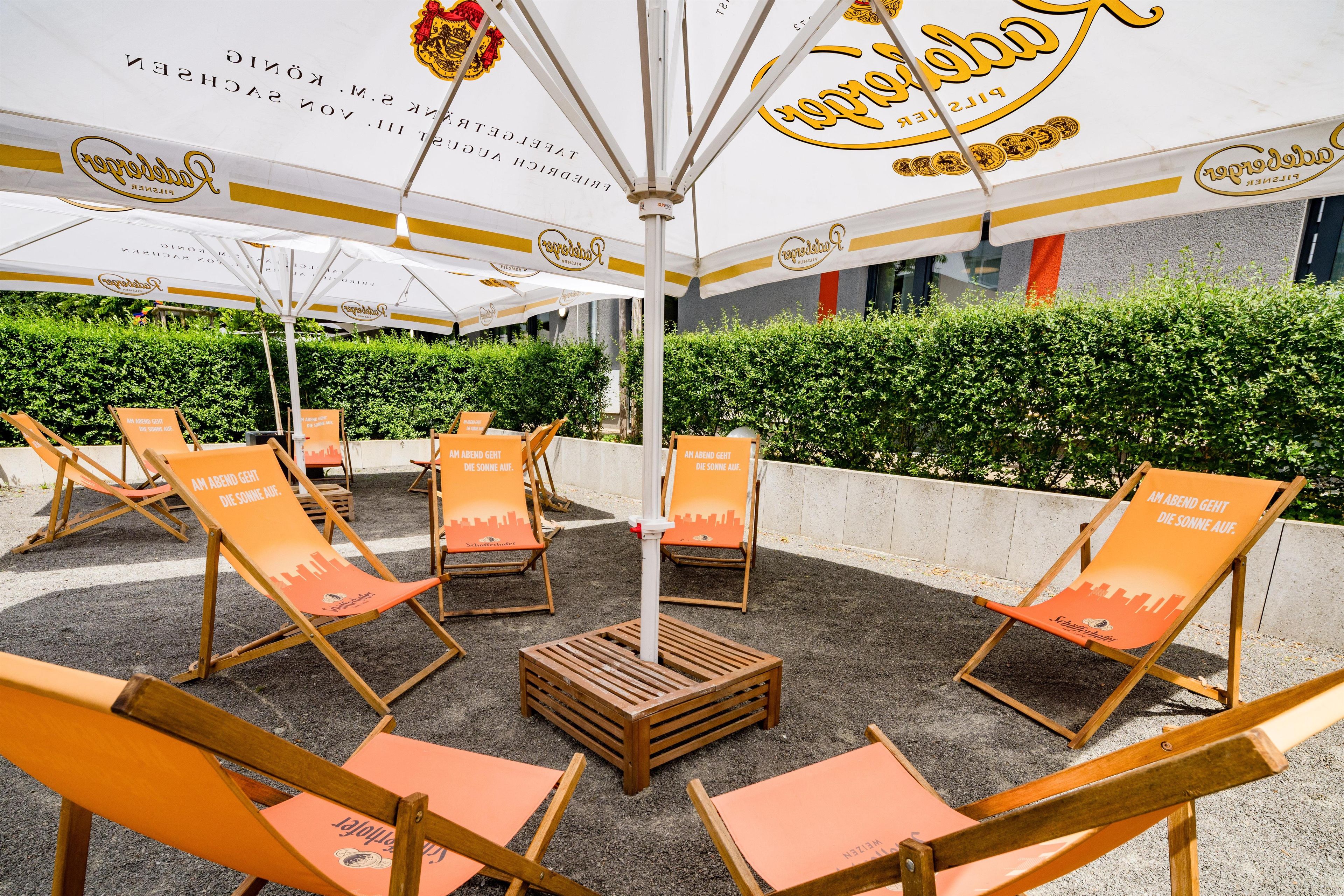 Outdoor seating with umbrellas, a wooden table, and a hedge in the background.