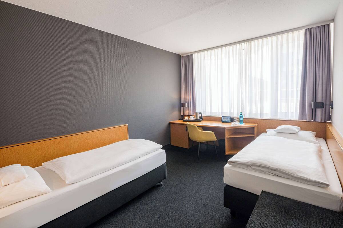 Une chambre d’hôtel avec deux lits, un bureau, une chaise et une grande fenêtre avec des rideaux.