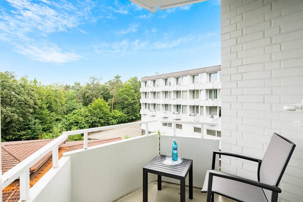 Un balcon avec une table et une chaise, une bouteille d’eau et une vue sur un immeuble et des arbres.