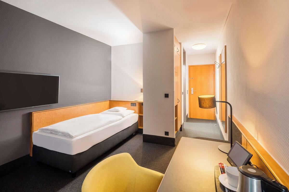 Une chambre d’hôtel avec un lit simple, un bureau avec un ordinateur portable, une chaise et un téléviseur fixé au mur.