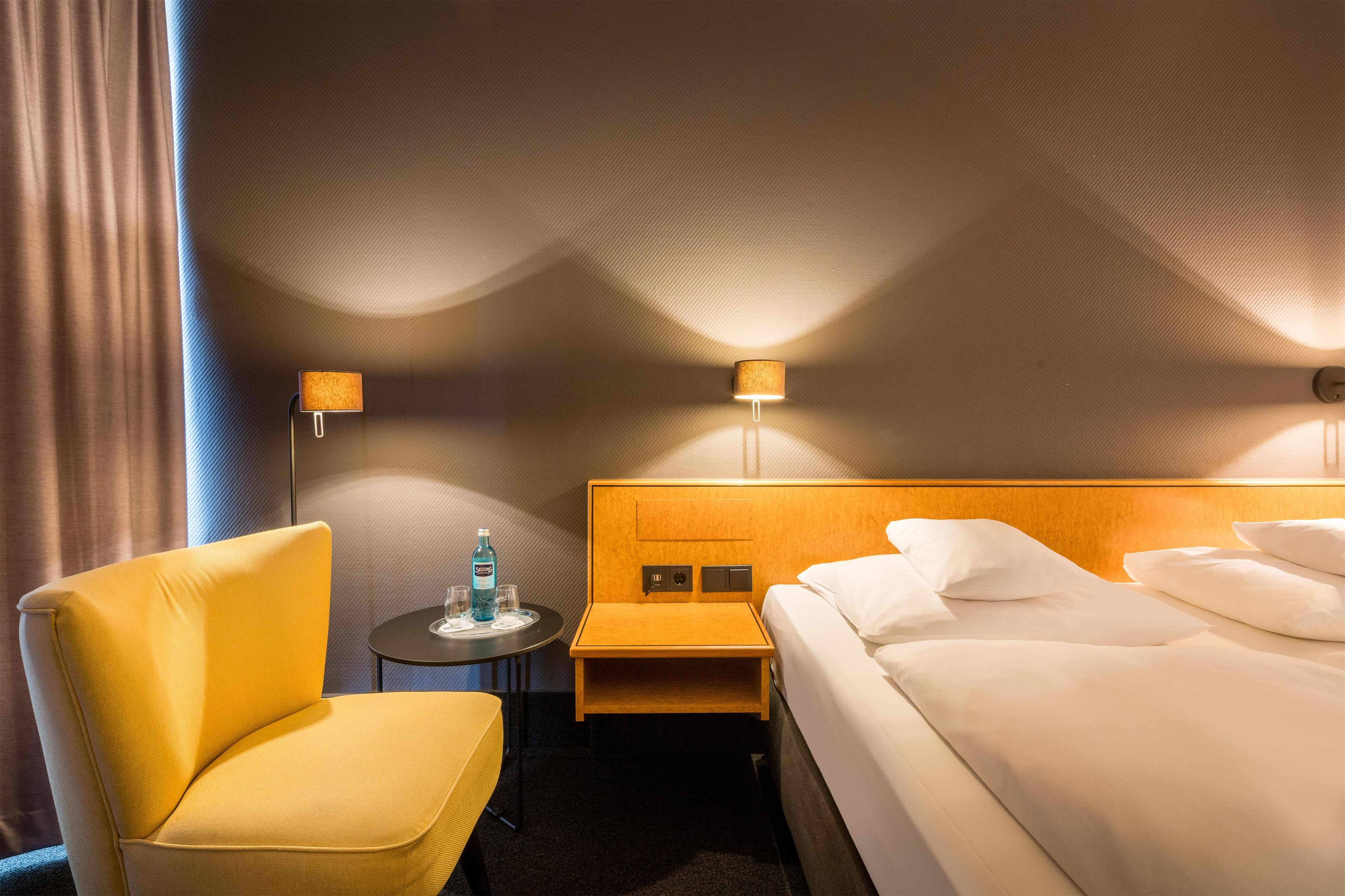 A hotel room with a single bed, a small table with a bottle and glasses, a chair, and two wall-mounted lamps.