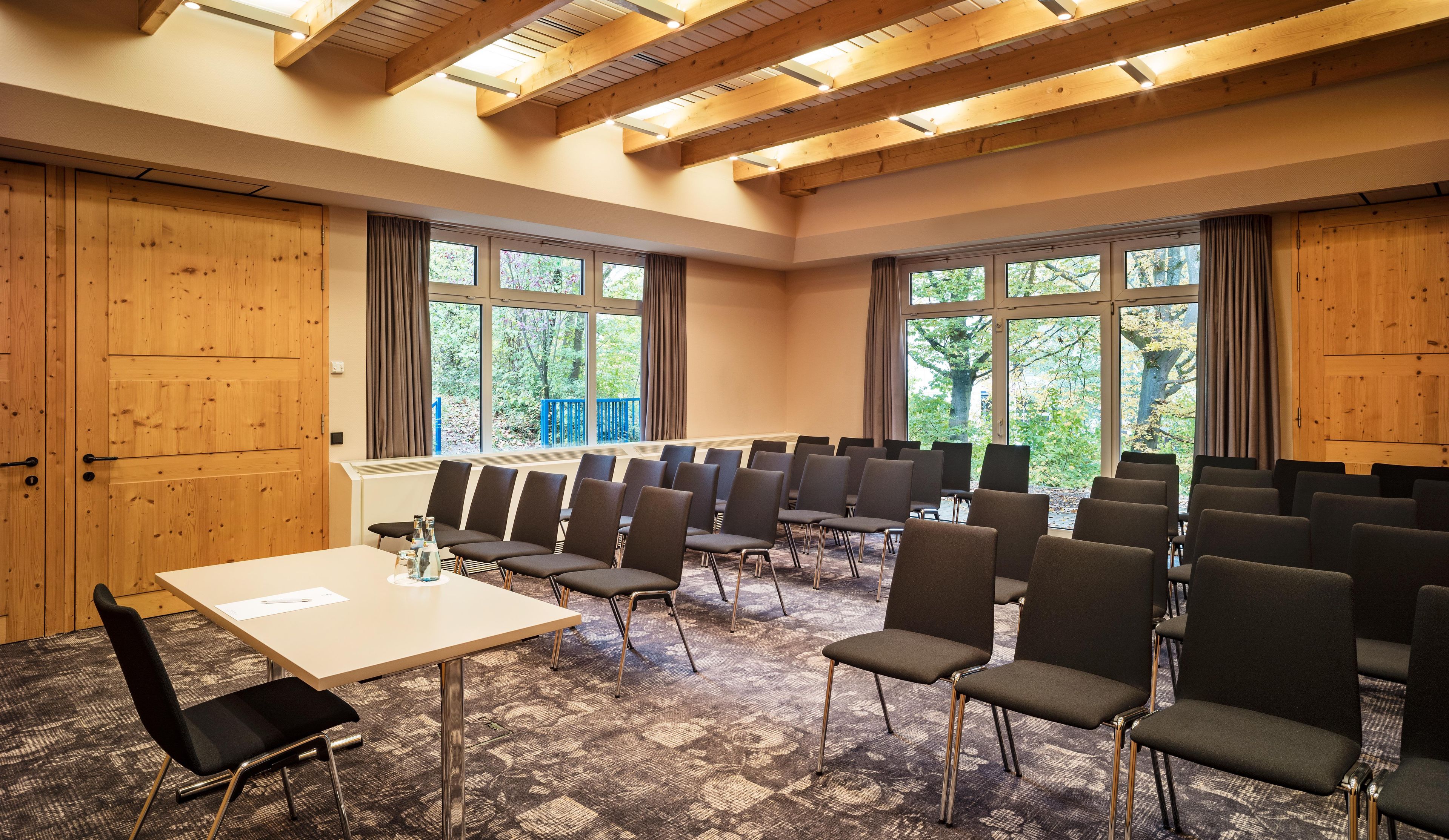 Ein Konferenzraum mit einem langen Tisch, mehreren Stühlen, Holzwänden, großen Fenstern und einem gemusterten Teppich.