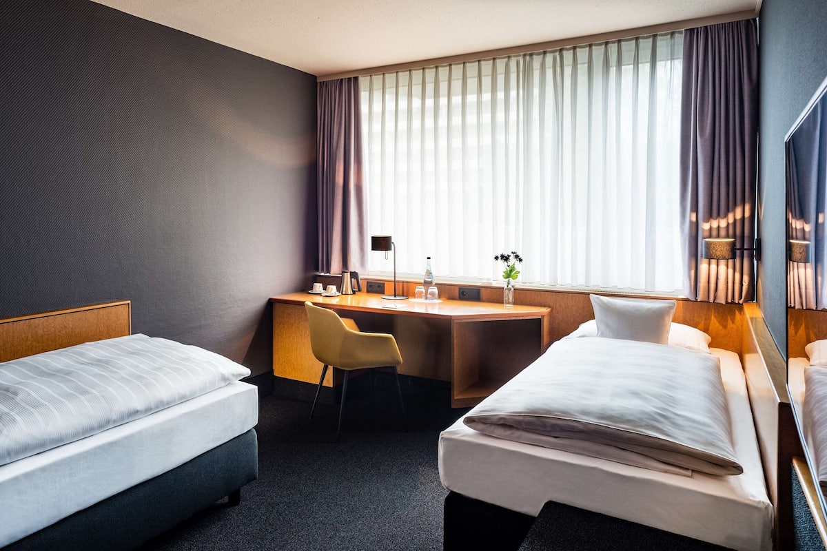 A hotel room with two beds, a desk, a chair, a lamp, and a window with curtains.