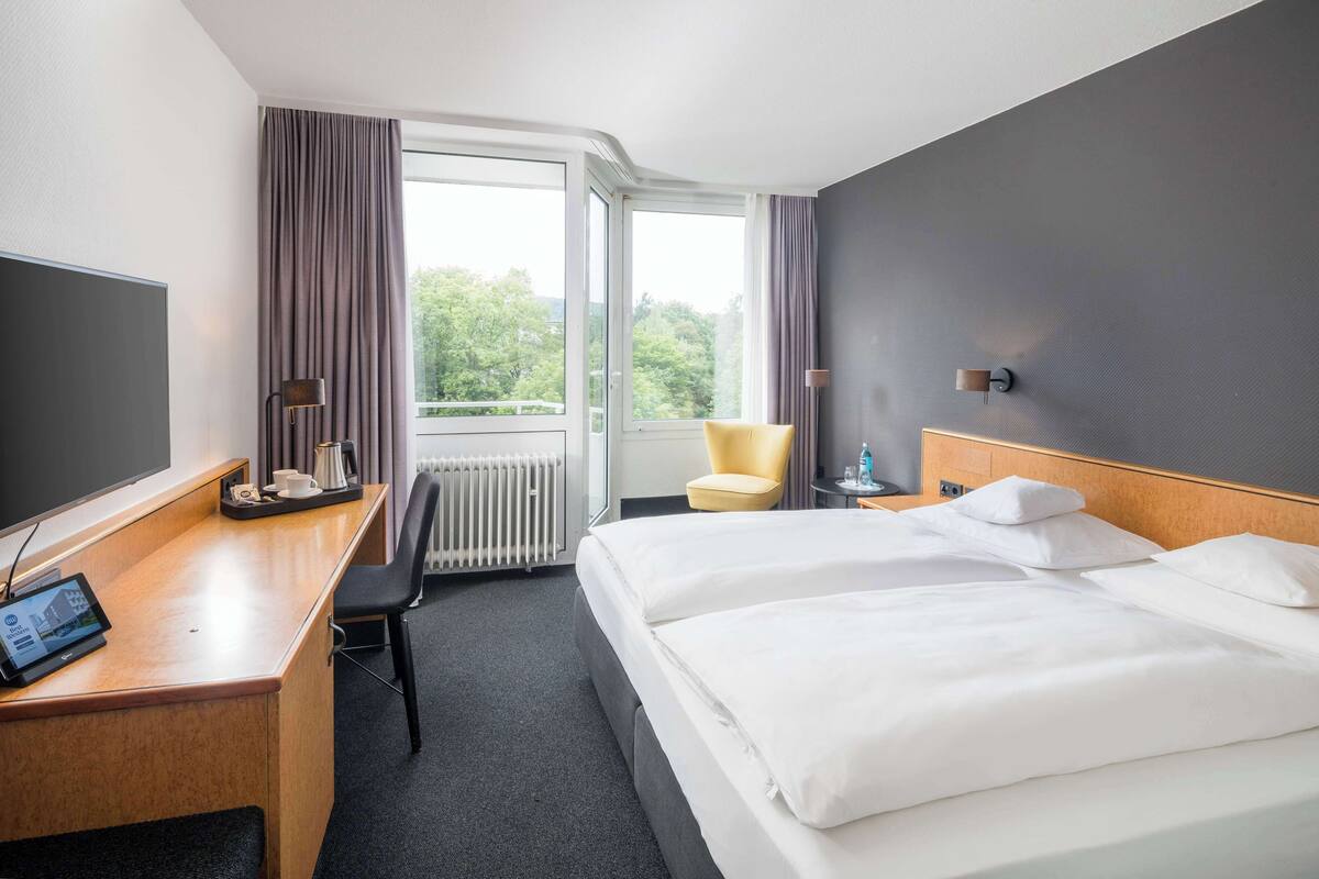 Une chambre d’hôtel avec un lit, un bureau avec un ordinateur, une chaise et une fenêtre avec des rideaux.