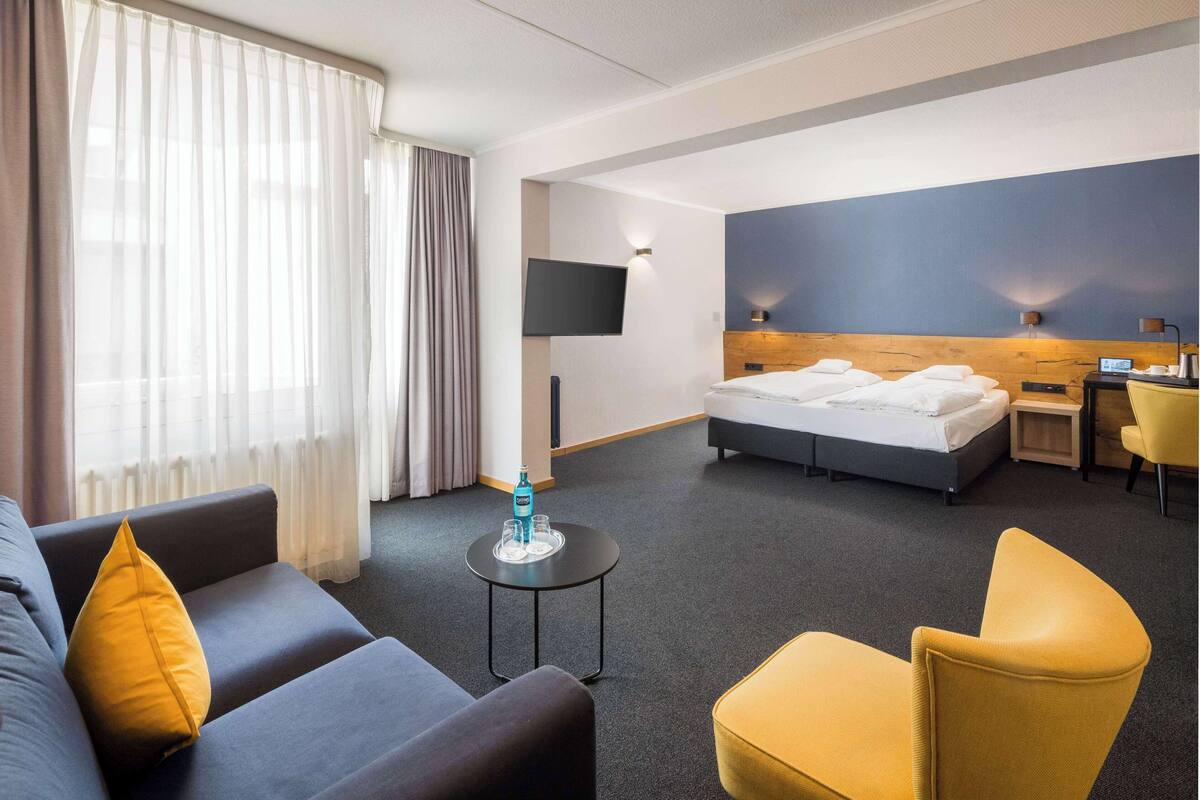 Une chambre d’hôtel avec un lit, un canapé, une chaise, une télévision et une petite table sur laquelle se trouvent une bouteille et des verres.