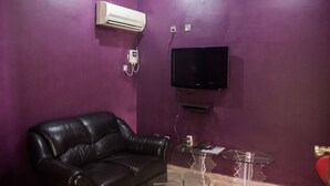 A room with a black leather sofa, a glass table, a red chair, a mounted TV, and an air conditioning unit on the wall. - De Prince Guest House (Lagos)