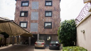 A multi-story building with a tiled facade, two cars parked in front, and a large umbrella providing shade. - De Prince Guest House (Lagos)