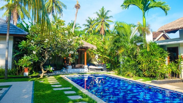 A pool surrounded by lush greenery and palm trees, with a thatched-roof structure nearby. - Villa Bau Nyale (Kuta)