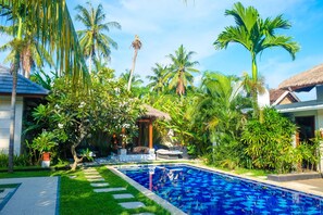 Una piscina rodeada de abundante vegetación y palmeras, con una estructura de techo de paja cerca.