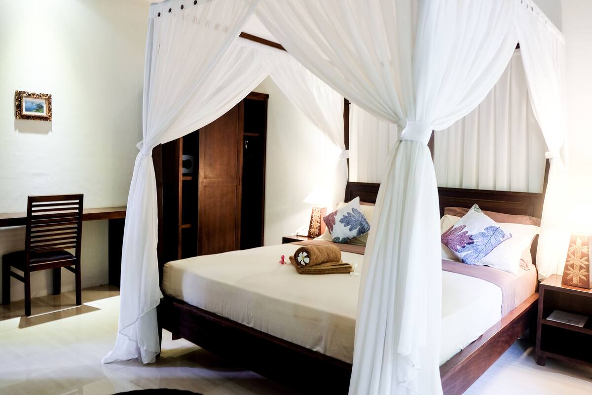 A four-poster canopy bed with a wooden headboard, a nightstand, a chair, and a framed picture on the wall.