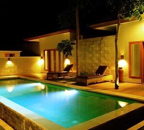 Una piscina con dos sillas reclinables y una mesita, rodeada de muros con un diseño texturizado e iluminación cálida.