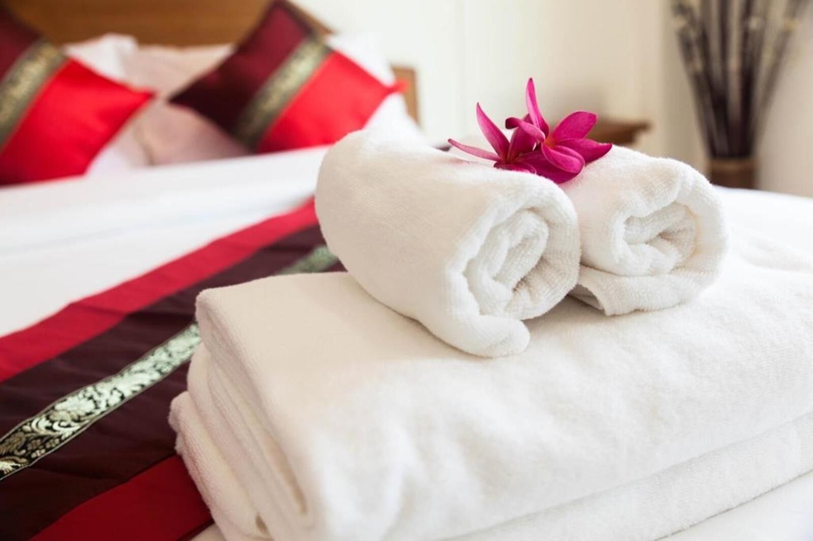 A neatly made bed with white and red bedding, a rolled white towel, and a pink flower.
