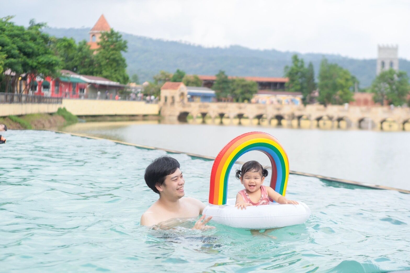 A man and a child in a swimming pool with a rainbow floatation device.