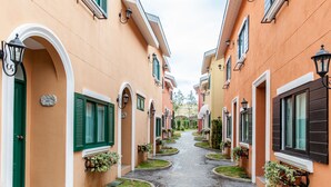 A narrow, paved pathway between buildings with green shutters and outdoor lighting fixtures.