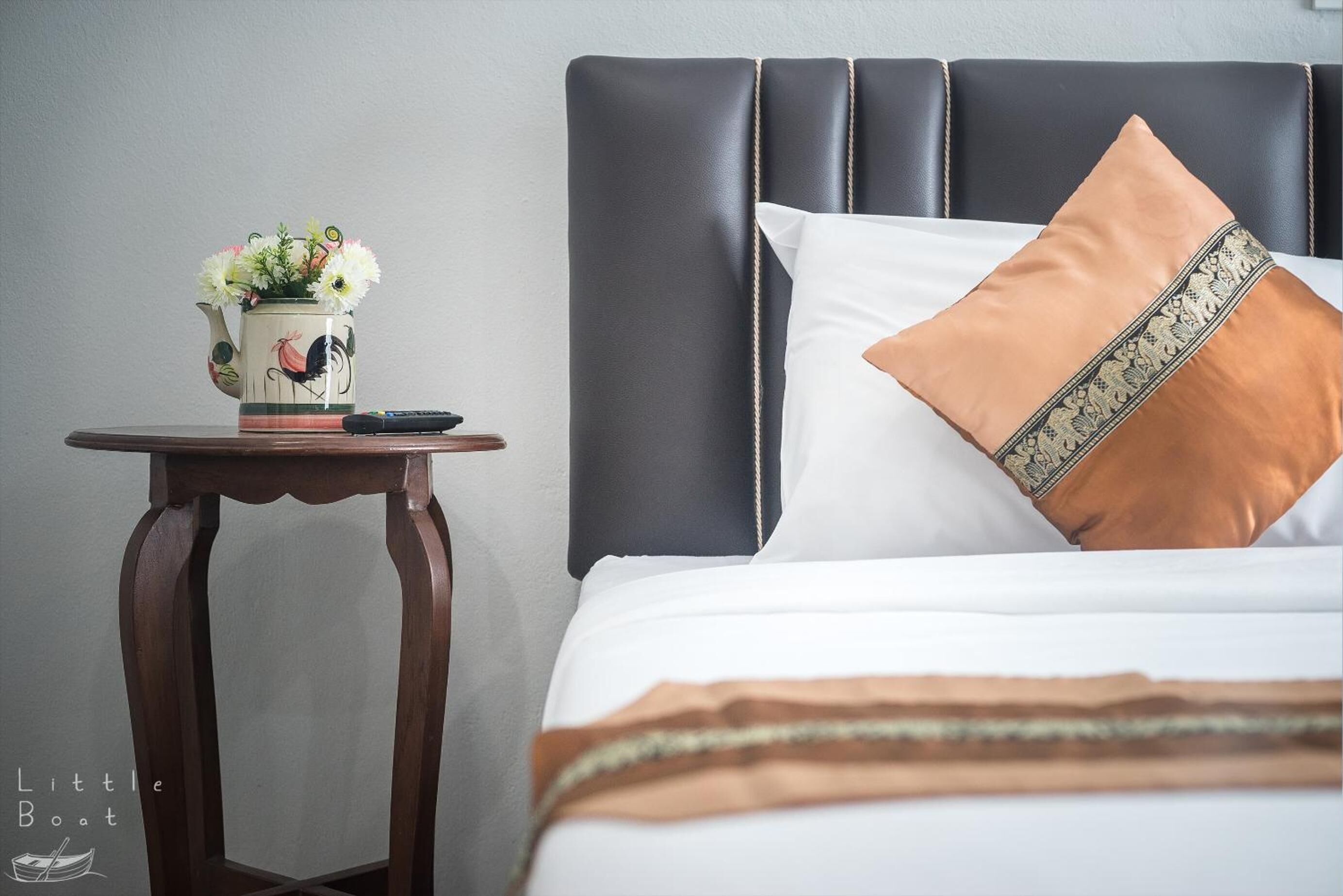 A neatly made bed with a white and brown pillow, a decorative vase with flowers, and a remote control on a wooden nightstand.