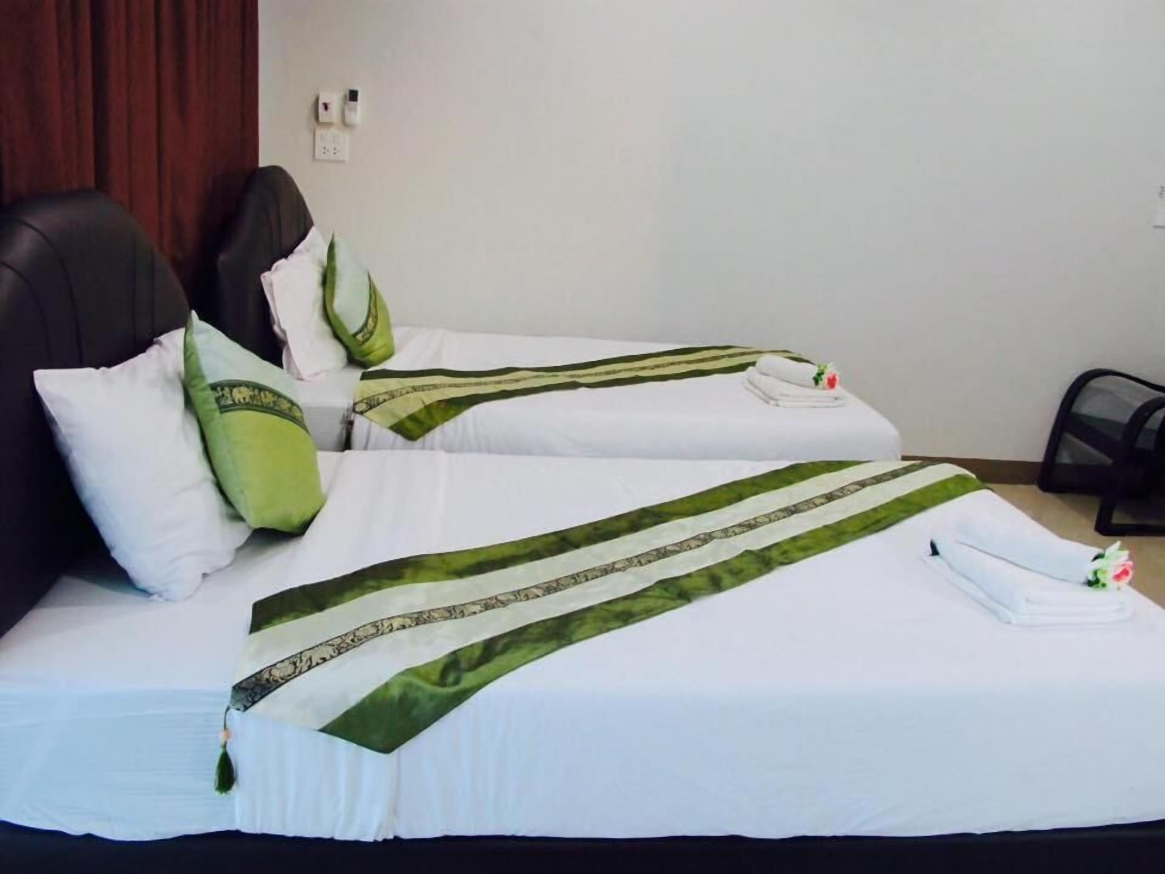 A hotel room with two beds, each with white bedding and green accents. There are pillows and a folded towel on the beds. A black chair and a small table are visible in the room.