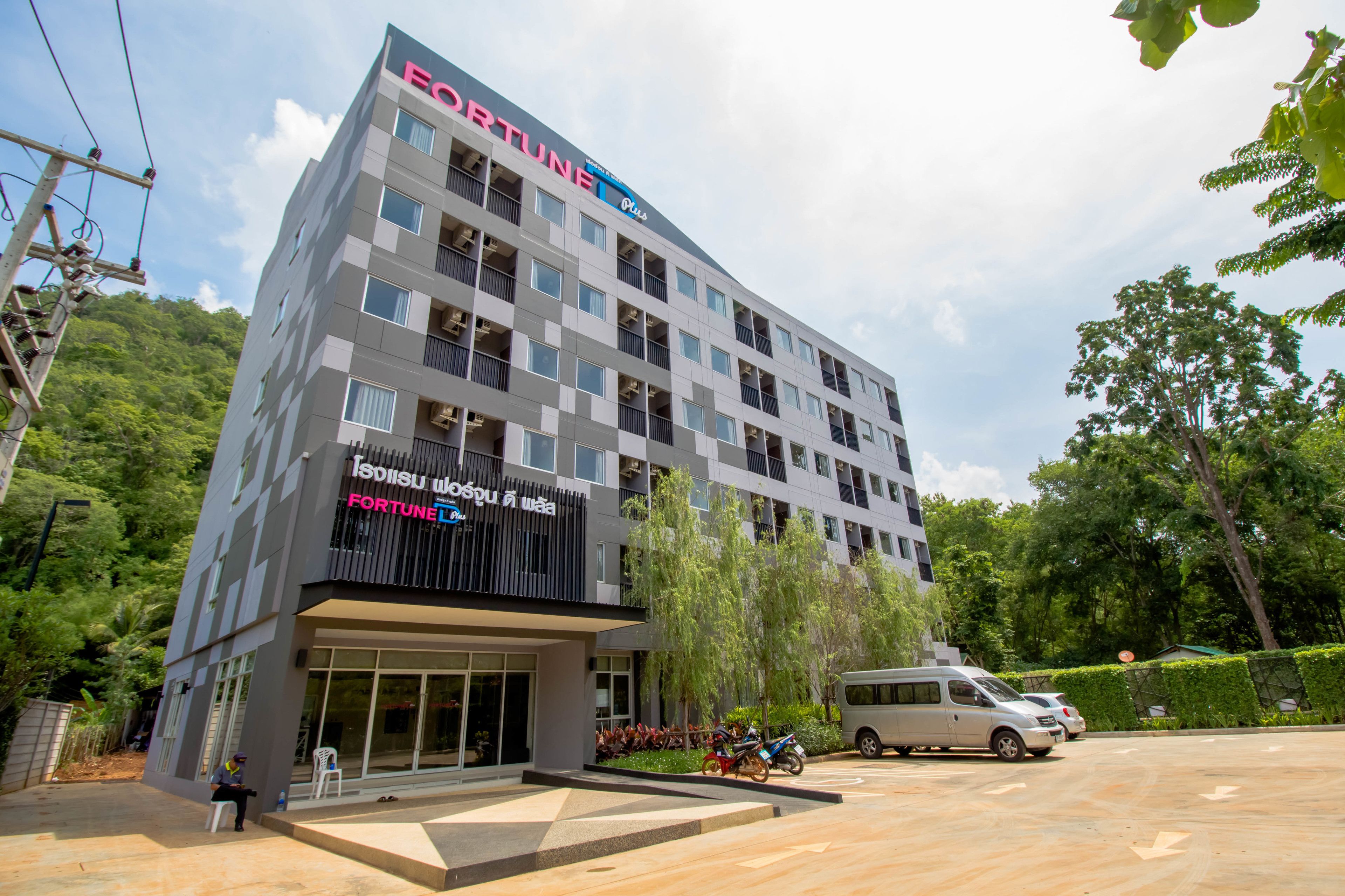 A multi-story building with a prominent Fortune hotel sign, surrounded by greenery and parked vehicles.