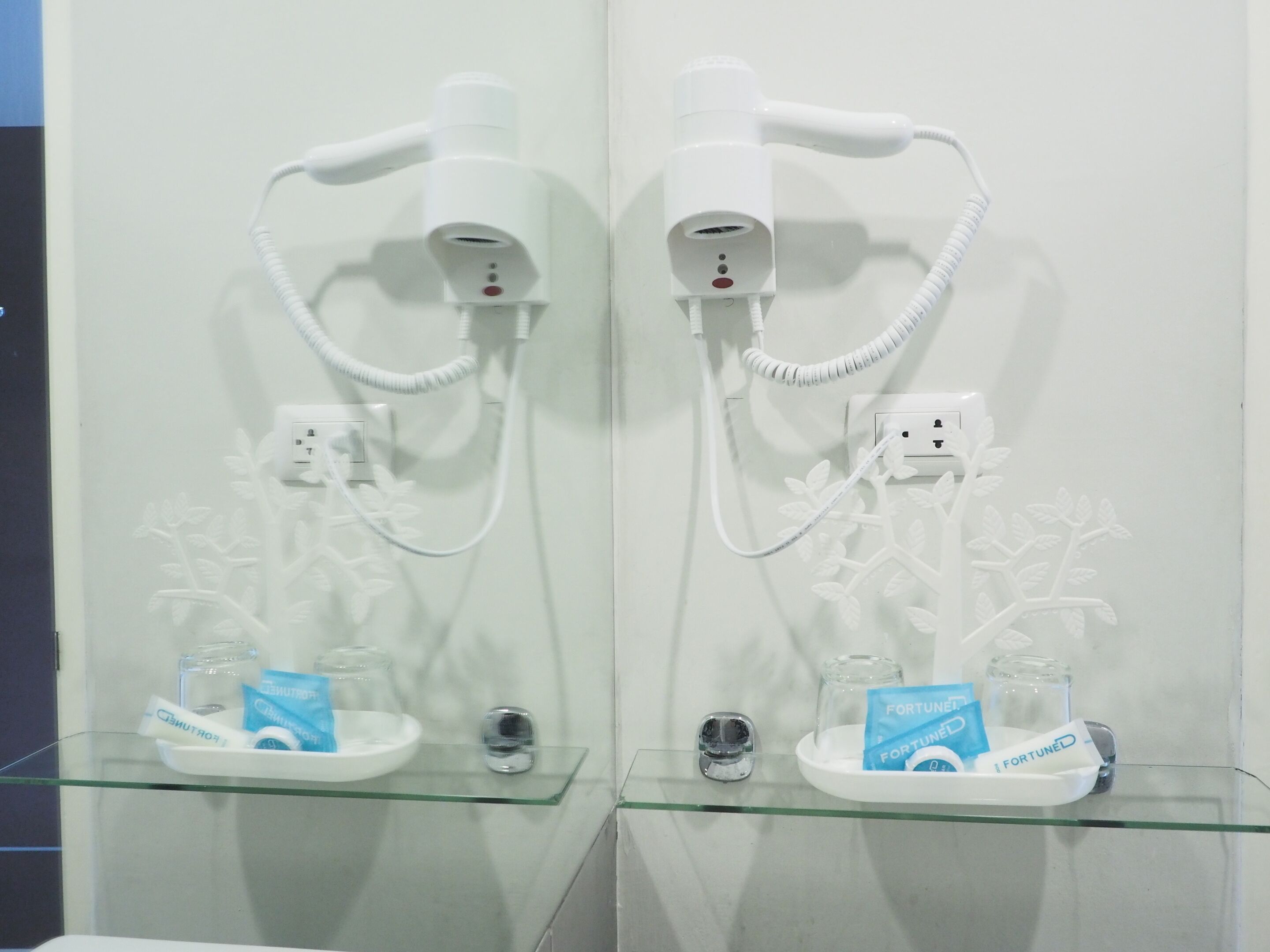 A bathroom with two hair dryers mounted on the wall, connected to power outlets. There are glass shelves with toiletries and a pack of toiletries labeled "FORTUNE."