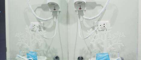 A bathroom with two hair dryers mounted on the wall, connected to power outlets. There are glass shelves with toiletries and a pack of toiletries labeled "FORTUNE."