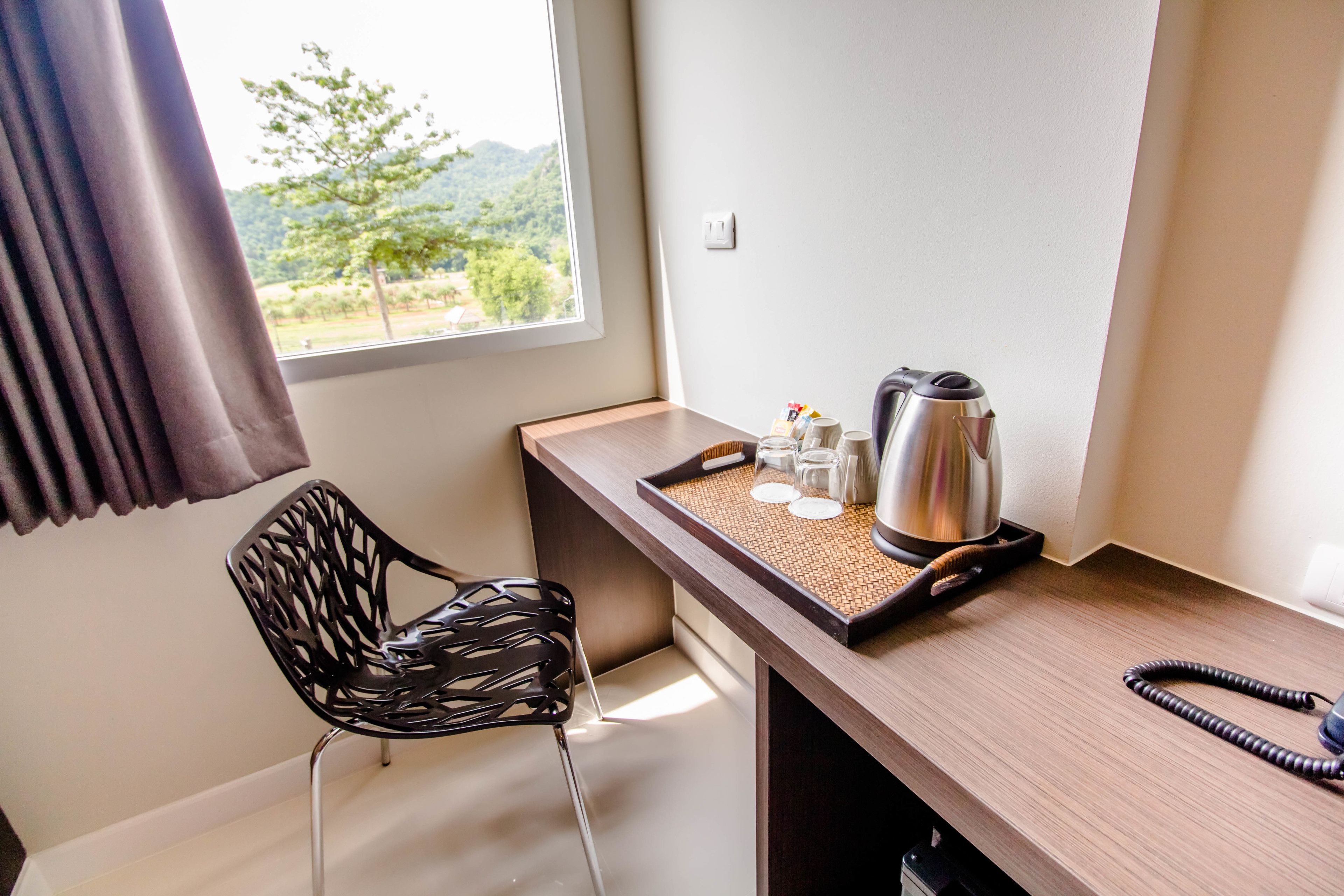 Een kleine kamer met een houten bureau, een zwarte stoel, een raam met uitzicht op groen en een waterkoker op het bureau.
