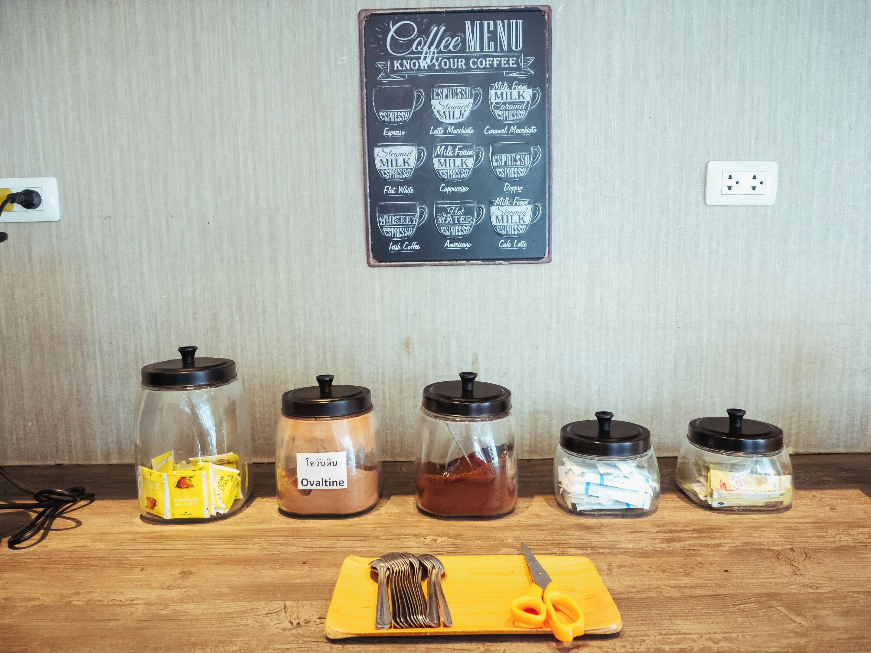 Un menu de café affiché sur un mur, différents types de grains de café dans des bocaux et une planche à découper avec des ustensiles.