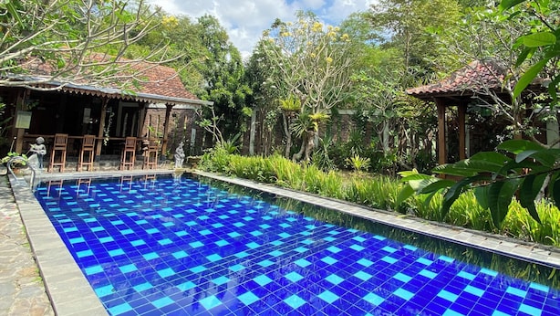 Piscina com azulejos azuis, rodeada por vegetação e duas pequenas construções com telhados em azulejo.