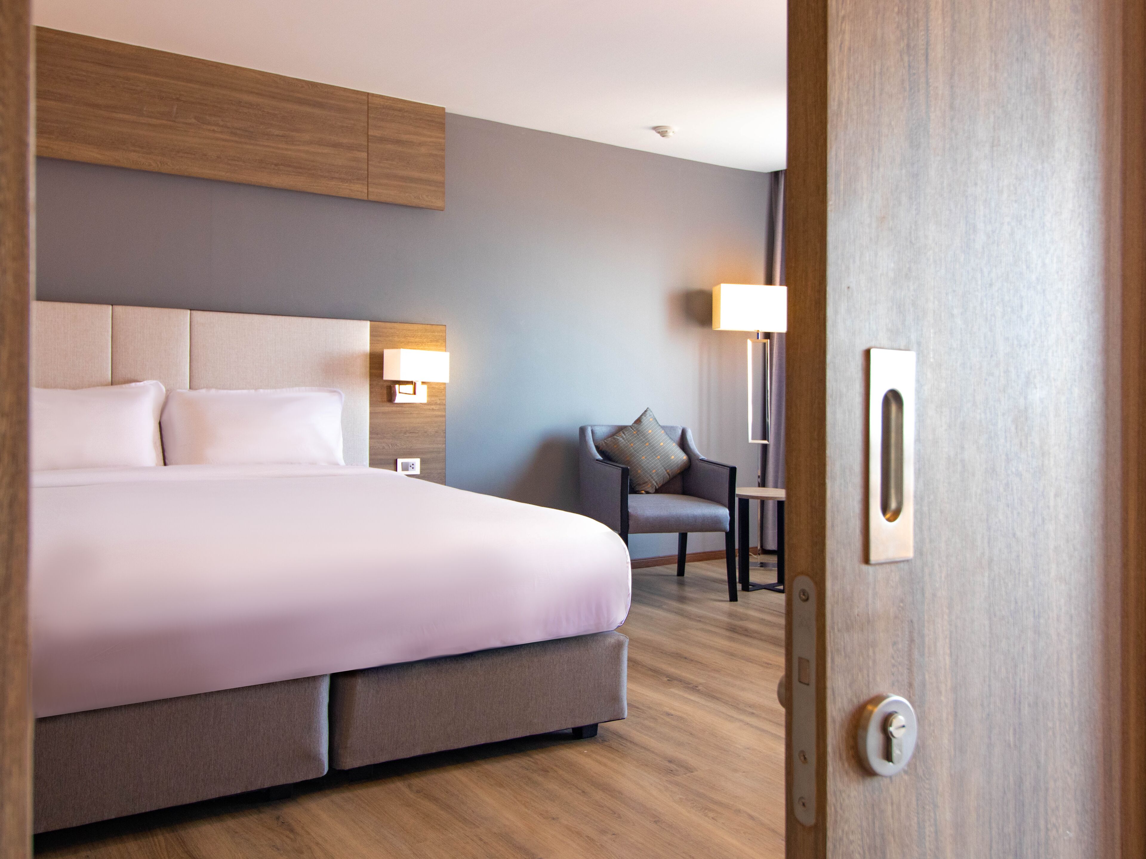 A modern hotel room with a large bed, a chair, a desk, and a lamp.