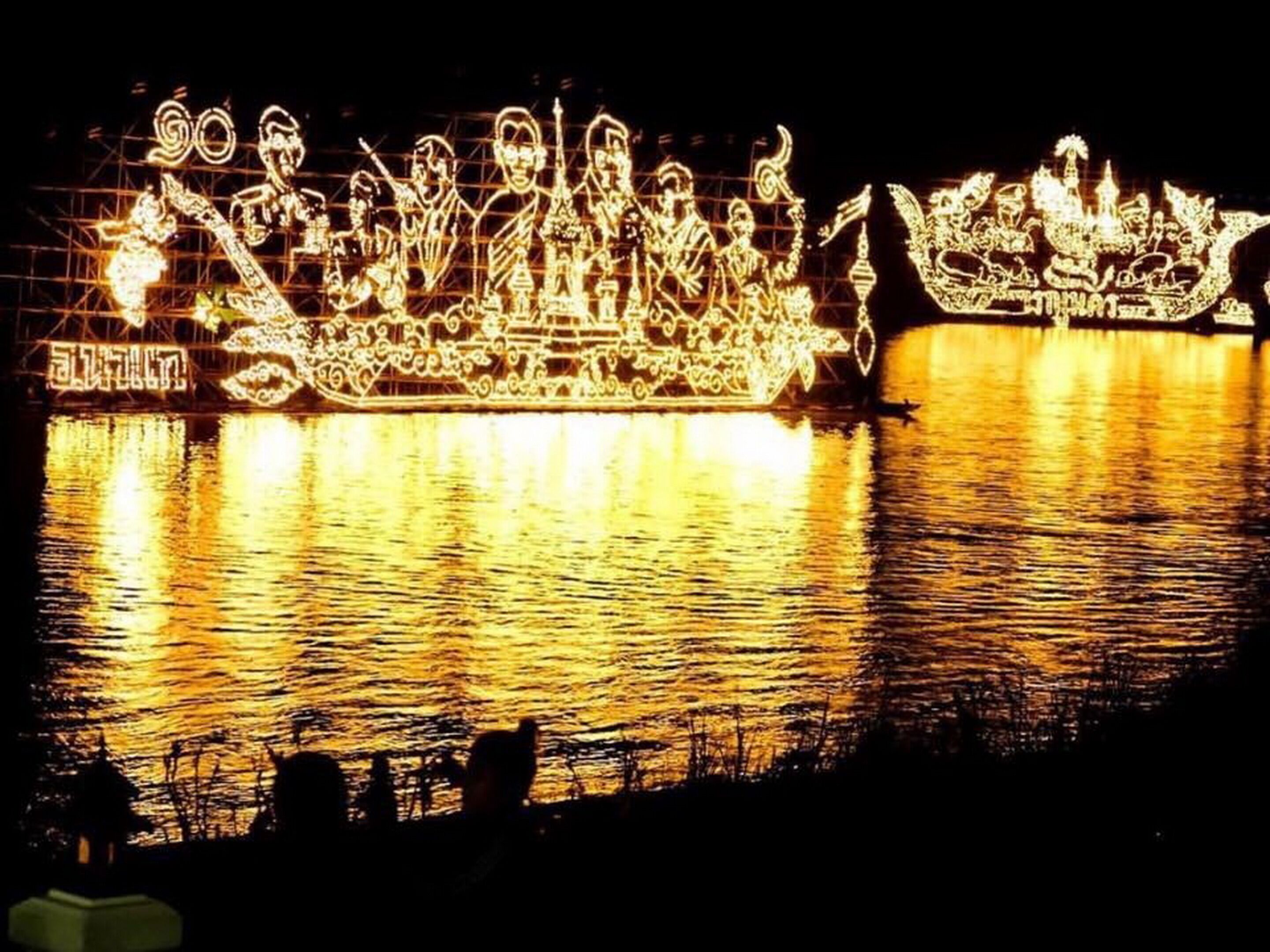 A nighttime view of a river with illuminated boat sculptures reflecting on the water.