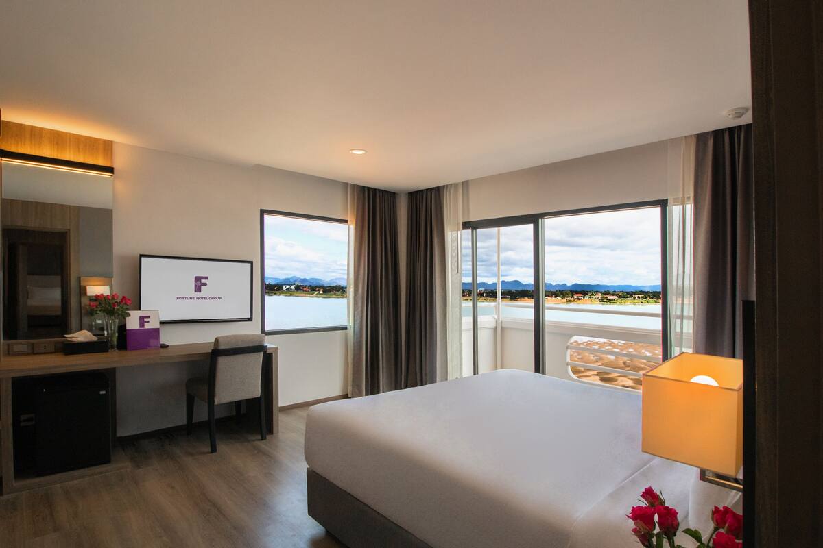 A hotel room with a bed, desk, chair, and large windows offering a view of a lake and mountains.
