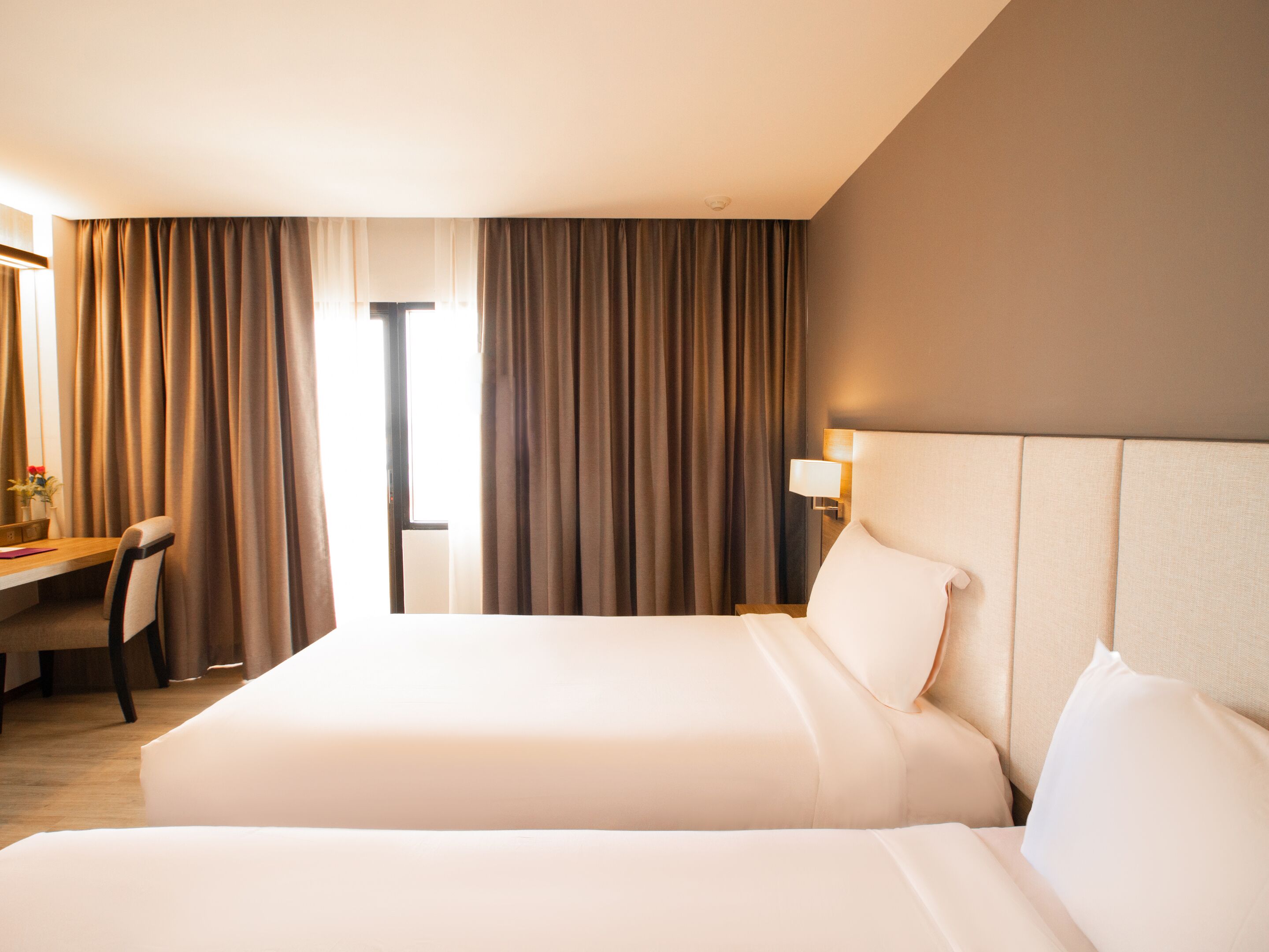 Ein Hotelzimmer mit einem großen Bett, einem Schreibtisch und einem Stuhl. Es gibt ein Fenster mit Vorhängen, eine Wandlampe und einen Blick nach draußen.