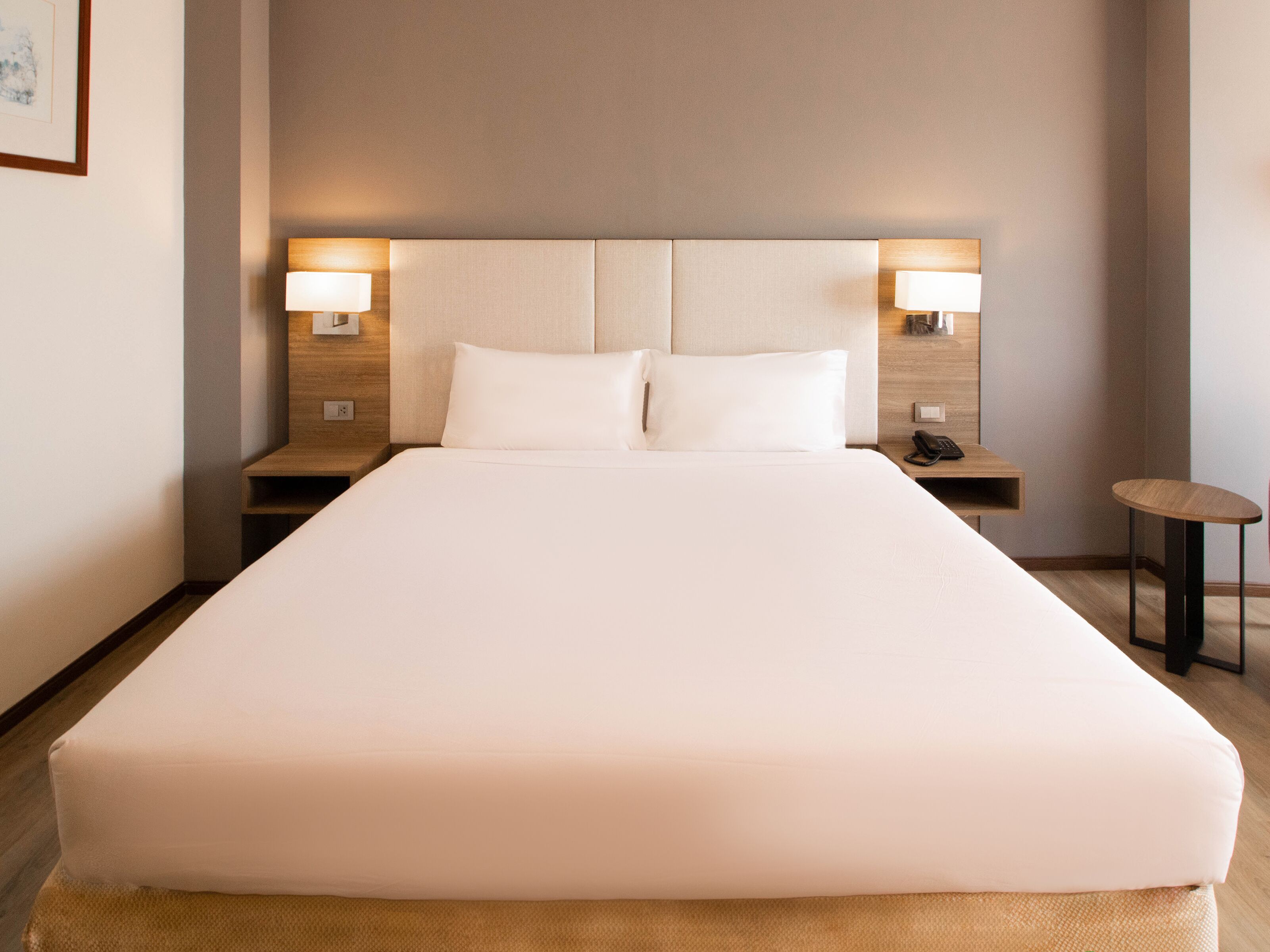A hotel room with a large bed, two bedside lamps, a small table, and a nightstand with a phone.