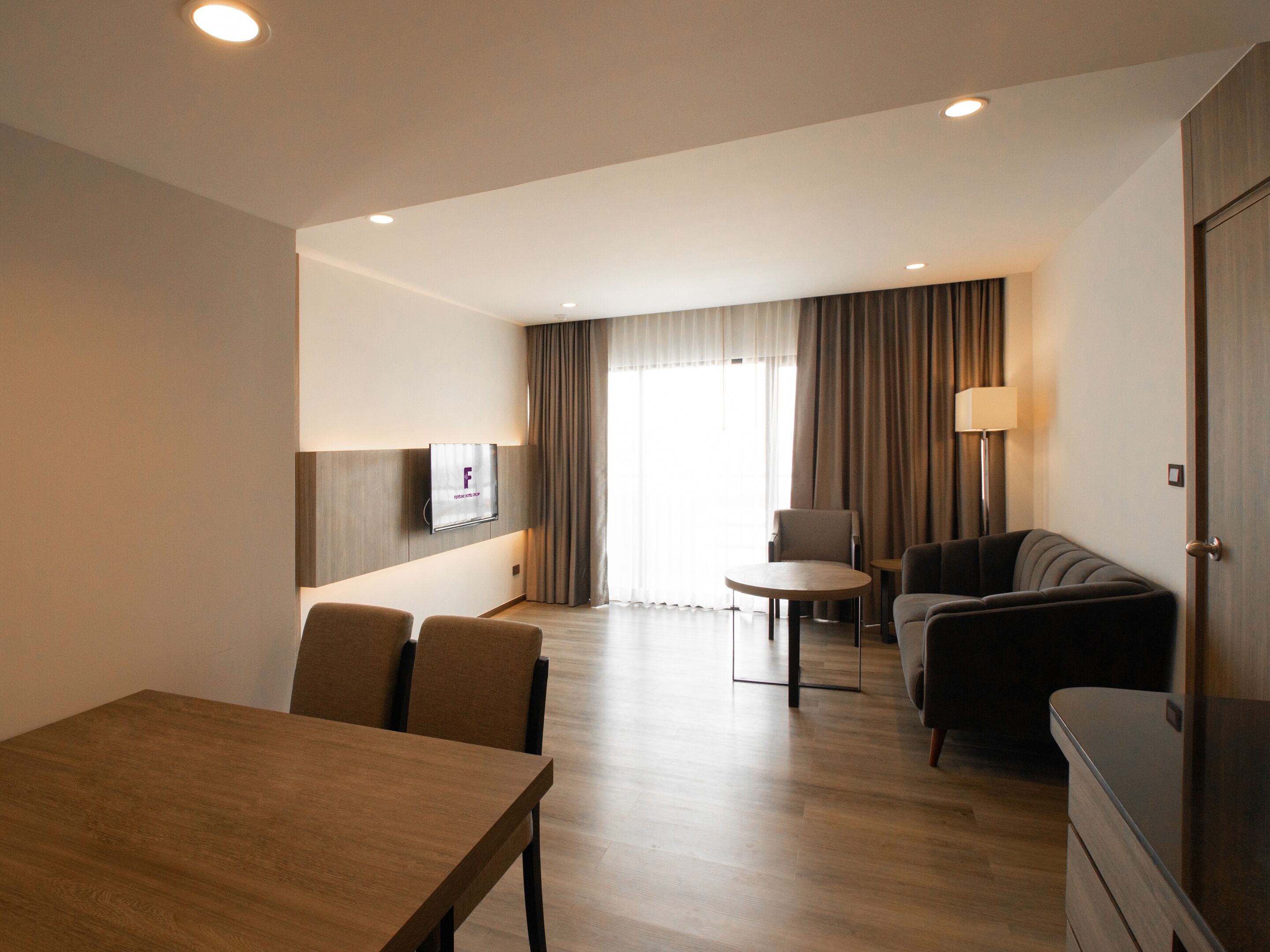 A modern hotel room with a dining area, a sofa, a TV mounted on the wall, and a large window with curtains.
