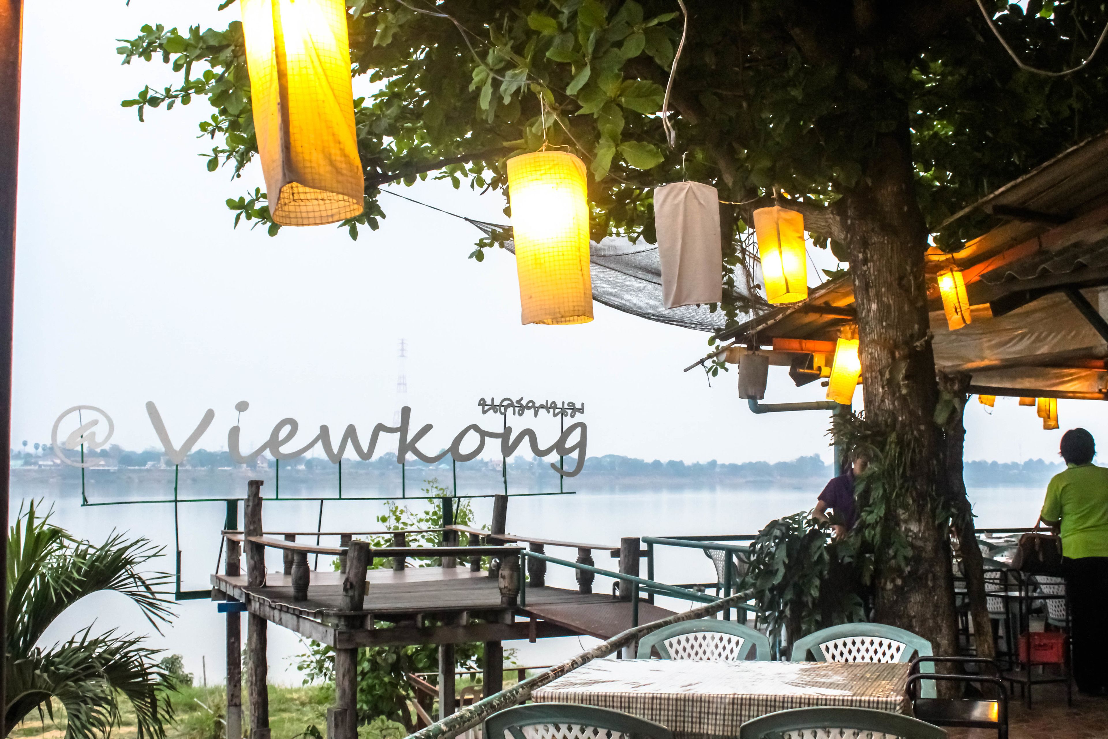 An outdoor dining area with wooden tables and chairs, hanging lanterns, and a view of a body of water.