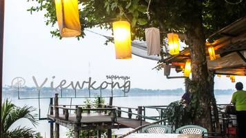 An outdoor dining area with wooden tables and chairs, hanging lanterns, and a view of a body of water.