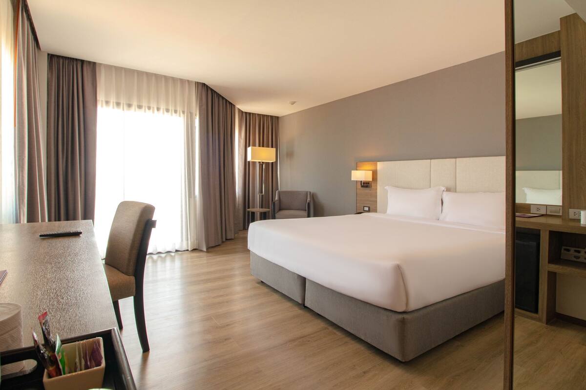 A hotel room with a large bed, a desk, and a chair. There is a lamp on the desk and a lamp on the wall.