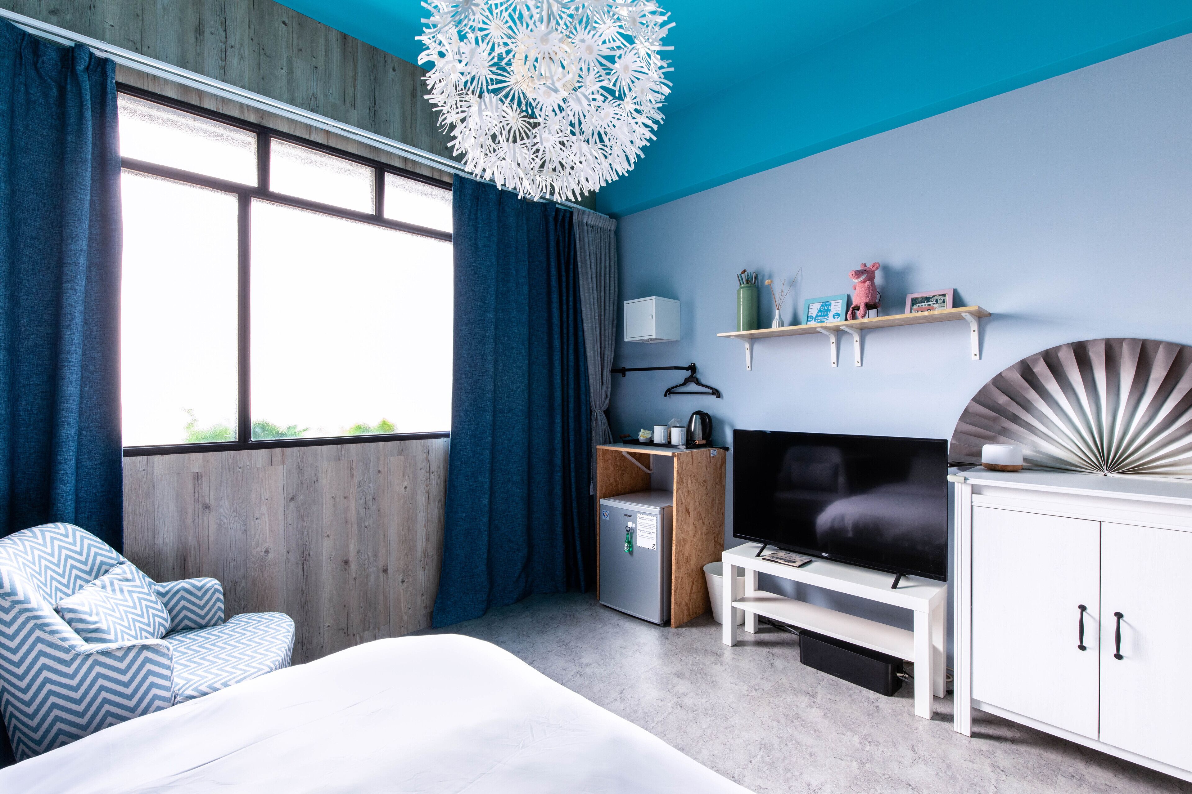 A modern hotel room with a bed, a TV, a small fridge, a wooden cabinet, and a decorative ceiling light.