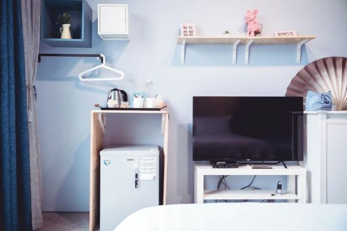 Hotelkamer met een bed, een televisie op een standaard, een kleine koelkast, een bureau met stoel en een plank met decoratieve objecten.