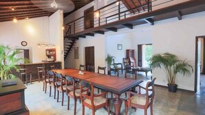 Un comedor amplio con una larga mesa de madera y numerosas sillas, una escalera que sube a un nivel superior y plantas en macetas.