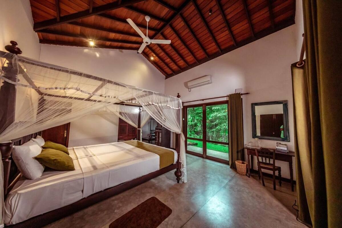 A bedroom with a canopy bed, a ceiling fan, a desk, and a window with a view of greenery.