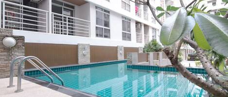 A swimming pool with a tiled deck, surrounded by a building with balconies and a stone wall.