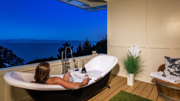 A bathtub with a view of the sea, a bottle, and a towel on a wooden deck. - Woodside Bay Chalets (Waiheke Island)