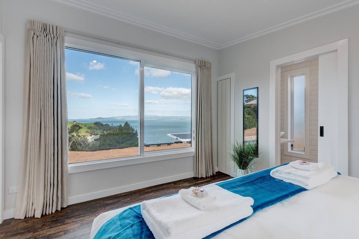A hotel room with a large window offering a view of the sea and greenery. There are two beds with white linens and blue towels.