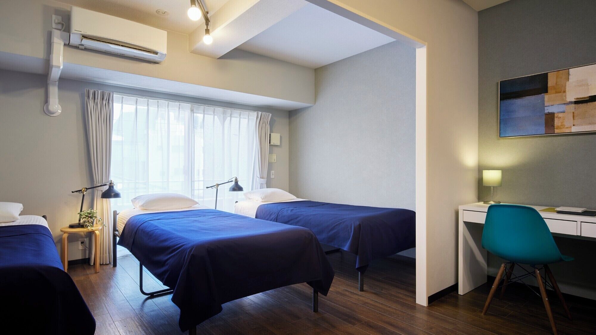 A hotel room with two single beds, a desk with a chair, a small table with a lamp, and an air conditioning unit.