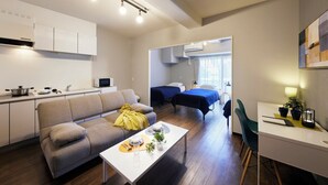 A modern apartment with a kitchen, a living area with a sofa and a dining table, two single beds, and a small desk area. - Minn Juso (Osaka)