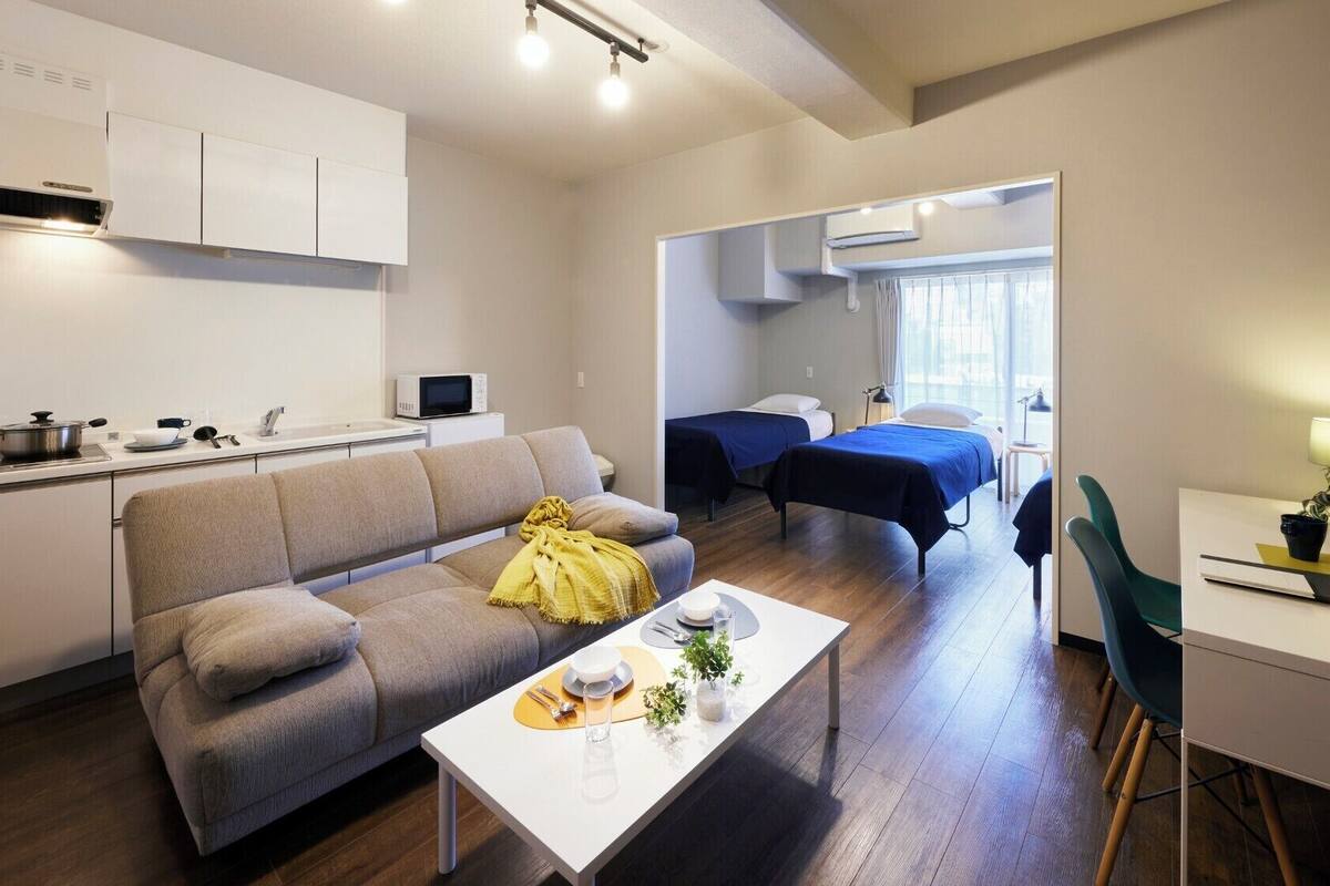 A modern apartment with a kitchen, a living area with a sofa and a dining table, two single beds, and a small desk area.