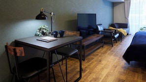 A room with a desk, two chairs, a bench, a TV, a bed, and a window with curtains.